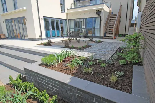 3 wide steps ascend to patio in pale paving next to garden walls capped with Steel Grey porcelain 20mm bullnose coping stones. 