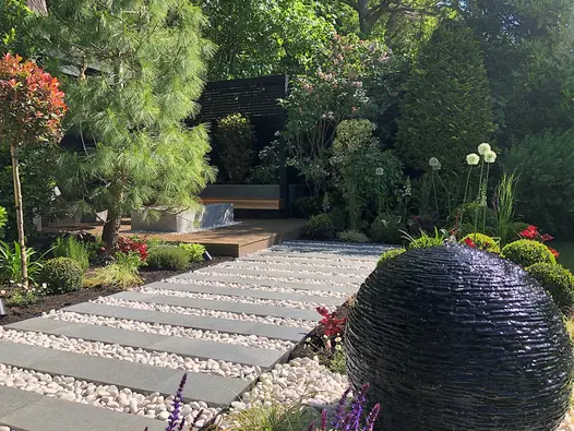 Egg-shaped slate water feature next to striped path of Black granite plank paving and white pebbles and beds with topiary.