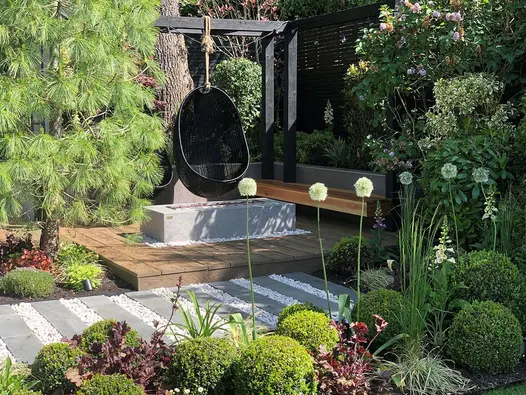 Black Granite plank paving and white pebble path runs past wooden deck with suspended egg chair. Design by Gardens of the Future.