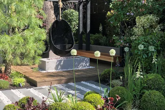 Black egg chairs hang from wooden pergola over vintage millboard decking, with firepit, bench and lush planting surrounding.