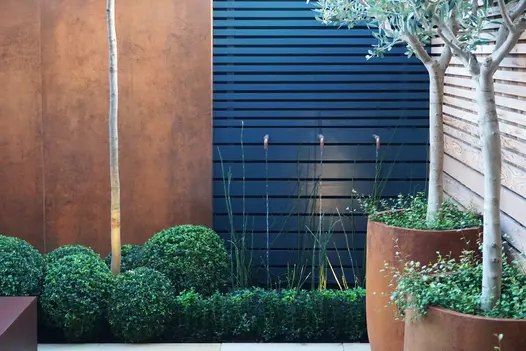 2 panels of steel corten designclad next to black fence has topiary bushes and potted trees lining the scape.