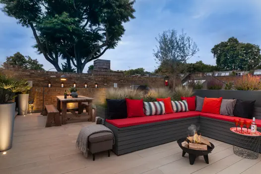 Small firepit burns in centre of corner bench on golden oak millboard decking surrounded by brick walls and garden lighting.