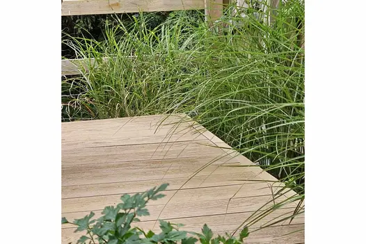 Long grasses creep out of corner of golden oak millboard decking space with wooden fence in the background.