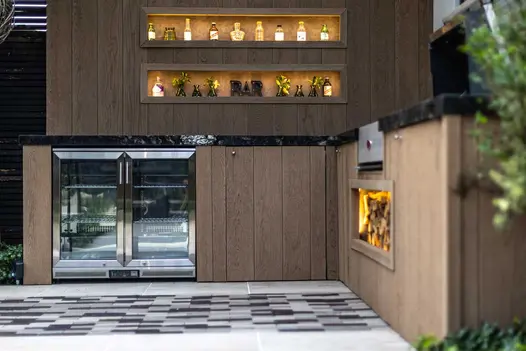 Corner of bar area adorned with bottles and small plants, clad in golden oak millboard and porcelain countertops.