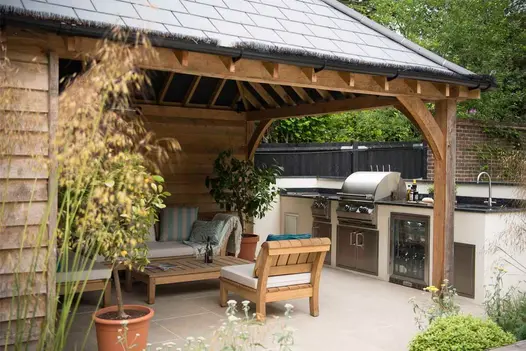 Roofed wooden-framed, open-sided room with wooden furniture abutting outdoor kitchen, paved in Golden Stone Porcelain.