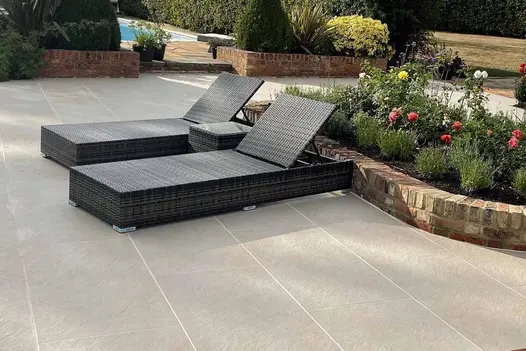 Two black rattan sunloungers sit on top of golden stone porcelain paving, with flowerbed behind and pool in the background.