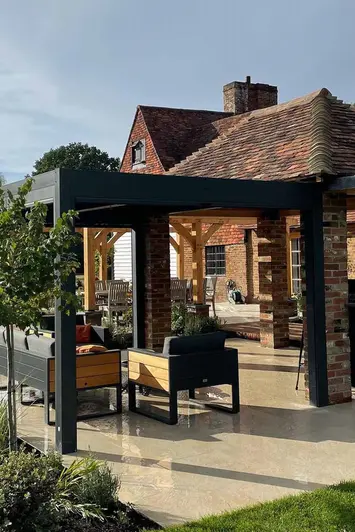 Garden furniture sits underneath dark metal pergola with wet golden stone porcelain paving showing off its texture.