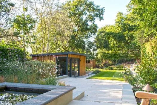 Looking across a heavily planted garden towards a cedar clad garden office adjacent to a lawn and Golden Stone porcelain patio.