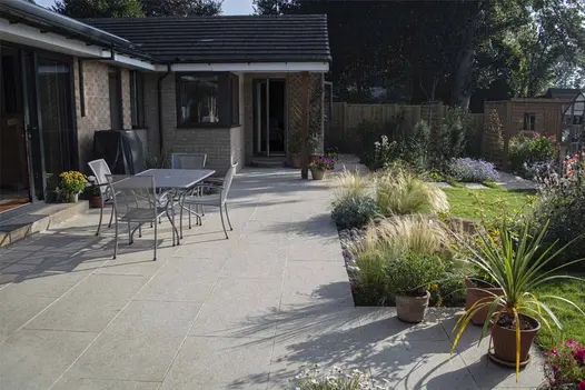Golden Stone Porcelain patio across width of rear of bungalow, with metal garden dining set and grassy planting in beds by Phil Hirst Garden Design.
