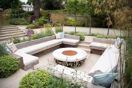 Square sunken seating area, large stone firebowl in centre, paved in Golden Stone Porcelain outdoor tiles, by Langdale Landscapes.
