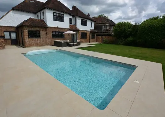 Rectangular swimming pool set in Golden Stone Porcelain paving surround next to lawn, behind 1930s suburban house.