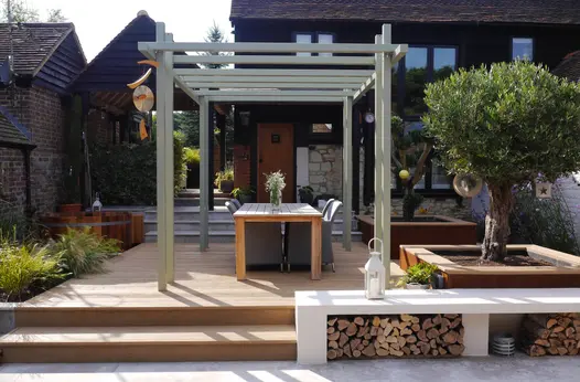 1 step leads up to golden oak millboard decking area with pergola and dining table, 2 olive trees in planters to the right.