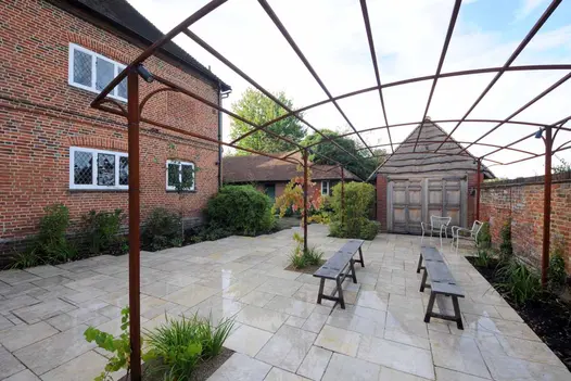 2 benches sit opposite each other under awning frame on Kota Brown limestone paving with gabled outhouse in background.