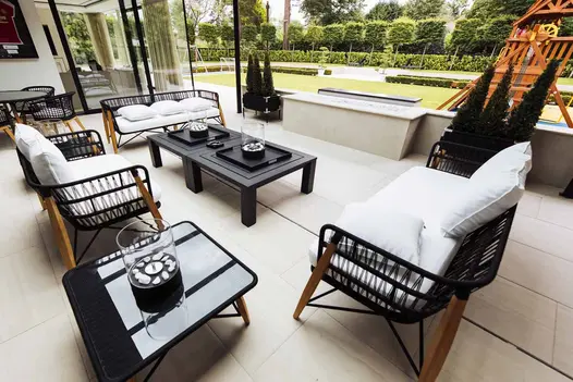 Patio of 900x600mm Faro porcelain paving with outdoor furniture low wall overlooking lawn and pleached trees.
