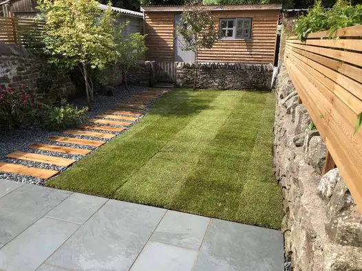 Patio with different widths of Brazilian Grey slate paving slabs fronts garden with lawn and plank path leading to garden office.