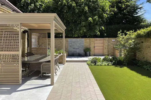 Path of Stone Grey brick pavers between pergola-covered dining area and fake lawn leads towards water feature in wall with Green slate cladding.
