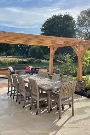 Ten seater wooden table and chairs sit under wooden pergola on a golden stone porcelain paving patio, lounge area at back.