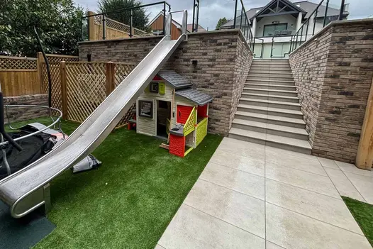 Play area with Grey York Porcelain Paving at the base of garden steps, featuring a slide and children’s playhouse.