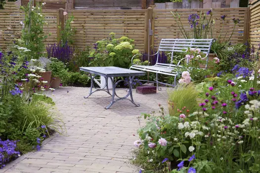 Metal garden furniture on Gromo Antica brick paving, with abundant green and purple foliage around against fence.