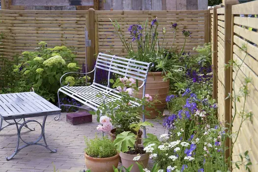 Metal garden furniture on Gromo Antica Clay Pavers, surrounded by green and purple planting against light coloured fence.