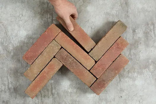 A hand adds 9th paver to 8 Seville brick pavers to 3rd row laid in zigzag pattern. Part of Chelmer Valley’s Alpha Collection.