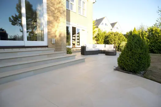Large patio of Harvest sawn sandstone, with 4 steps up to sliding doors, stretches across back of house. Shrubs in background.