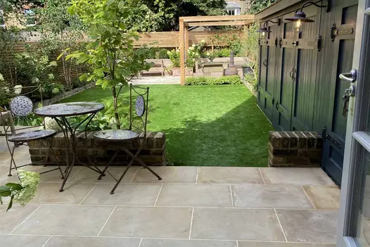 Long garden with bistro set on shady Harvest smooth sandstone patio, large square lawn and pergola-covered paved area beyond.