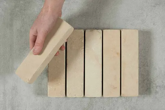 5 Harvest sandstone patio bricks in row on grey background. A hand is placing a 6th brick at an angle on top.