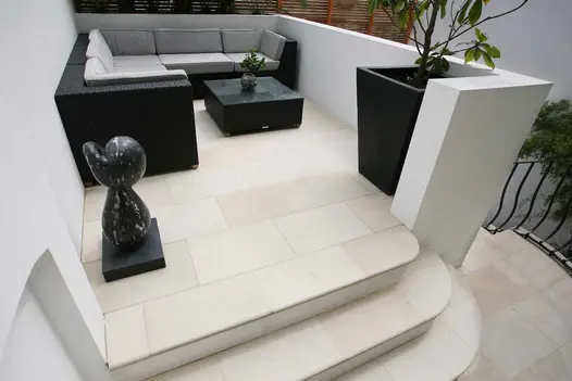 3 bullnose steps with curved corner rise to Beige smooth sandstone paving enclosed by white wall, with dark furniture.