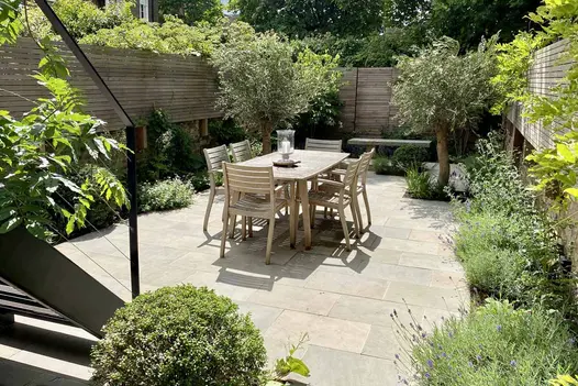 Metal steps descend to small oblong urban garden with planted borders edging Heath smooth sandstone paving.