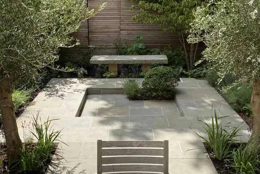 Paved area with square shallow sunken centre in fenced garden with stone bench and olive trees. 
