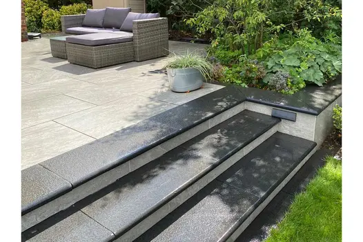 3 Black granite bullnose steps with pale risers ascend to porcelain-paved patio. Top step continues as coping of raised bed.