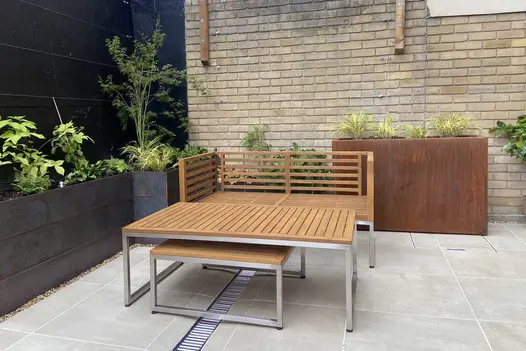 Modern bench and table in walled courtyard paved with Cream porcelain paving and channel drain. Tall planters around edge.