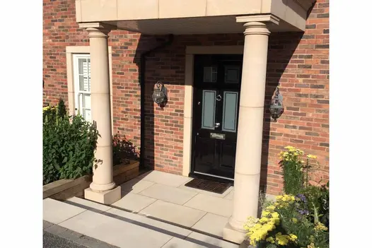 two steps down to front door utilising the beige smooth sandstone paving slabs with two large pillars holding porch either side of the door.