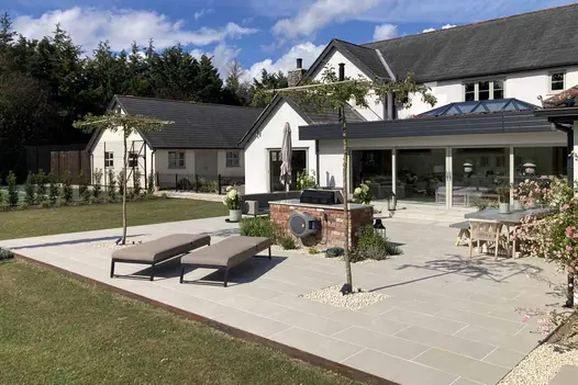 Slab Khaki porcelain patio with outdoor furniture, loungers and young trees outside huge glass doors of white gabled house.