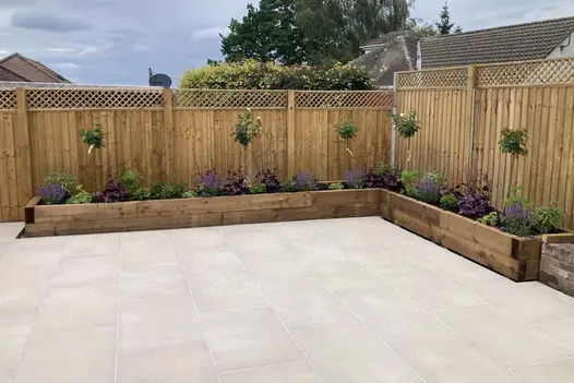 Corner of garden edged with tall fence and L-shaped wooden raised bed. Area completely paved with Venetian beige porcelain slabs.