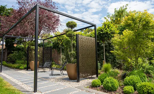 Plank paving slabs as stepping stones in black granite run down narrow path under pergola with 2 metal chairs sitting underneath.