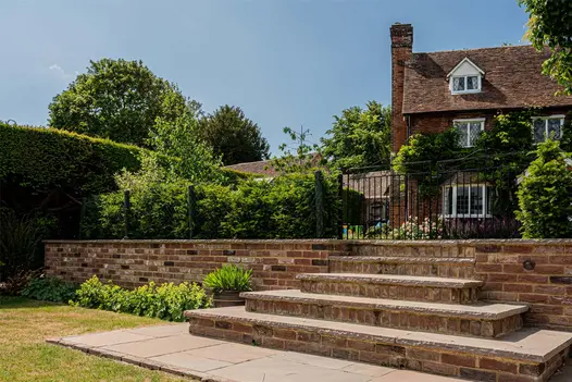 5 Autumn Brown sandstone steps up from paving laid in lawn to double iron gates in low hedge with rural cottage beyond. 