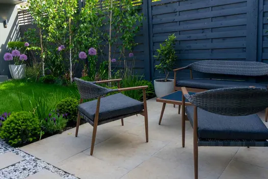 Blue-cushioned outdoor sofa and chairs sit next to matching fence on Jura Beige porcelain patio, next to planted border and lawn.