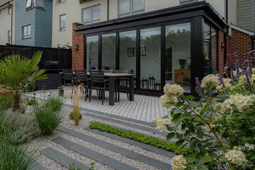 Charcoal Porcelain Planks used as stepping stone patio tiles leading up to back of house with glass conservatory and dining table.