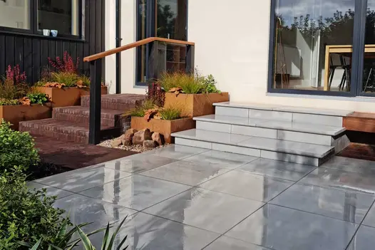 3 Dove grey bullnose steps rise from patio in matching paving to sliding patio doors, next to Corten steel planters.