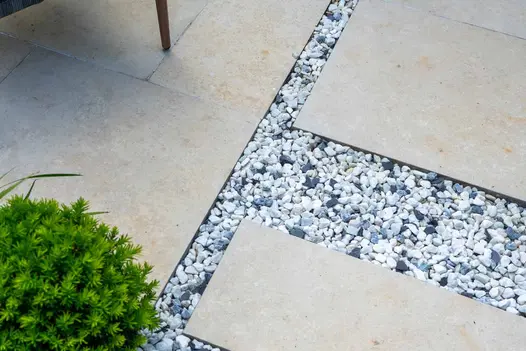 3 sections of Jura Beige porcelain paving separated by T-shape of blue and white pebbles. Small yew topiary in bottom left corner.