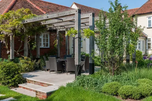 2 steps up to Jura Beige smooth sandstone patio with tall wooden pergola over dining set at back of extended cottage.