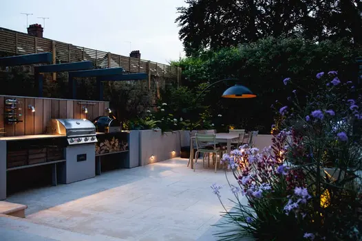 Twilight view of outdoor kitchen paved with Jura Beige sawn limestone with spotlights lit on splashbacks and raised beds.