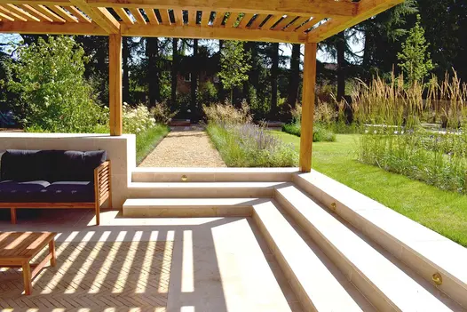 Seating area under pergola, paved with Jura Beige sawn limestone slabs, with 3 steps descending on 2 sides from lawn and path.