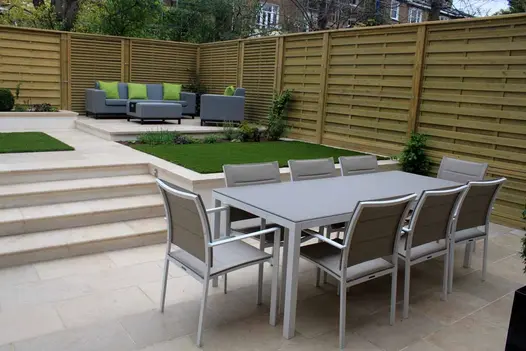 Fenced garden with Jura Beige sawn limestone patio and 4 steps rising to more paving that edges square patches of grass.