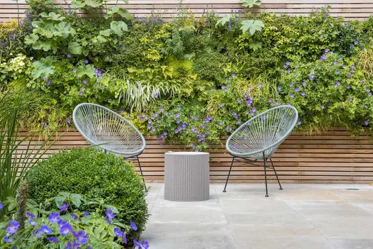 2 wire bucket chairs by small table on Jura Beige sawn limestone paving in front of raised bed faced with wood battens.
