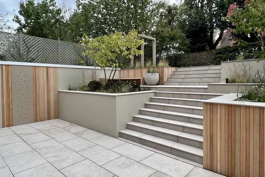 Deep beds retain steep slope on either side of wide flights of steps rising from area paved in Jura Grey porcelain outdoor tiles.