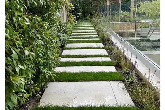Long, straight path of Jura Grey porcelain paving slabs, with wide grass joints, leads past glass railings overlooking paved area.