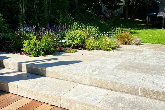 2 shallow steps of Jura Grey smooth limestone paving, with matching risers, ascend to paved area with planted border next to lawn.